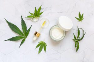 Pipette with oil and opened cream jar near green cannabis leaves